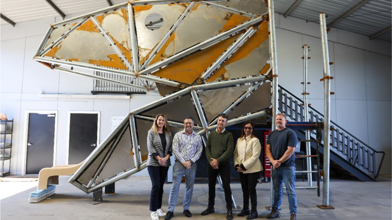 Group of five people standing in front of a large geometric metal sculpture inside an industrial workshop or fabrication facility