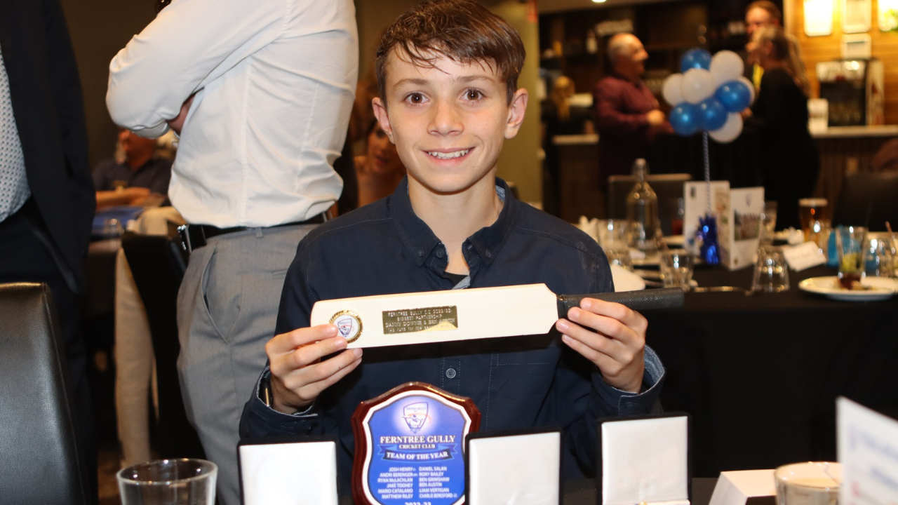 Ben Austin holding a miniature cricket bat trophy at the Ferntree Gully Cricket Club awards night, with plaques and decorations on the table