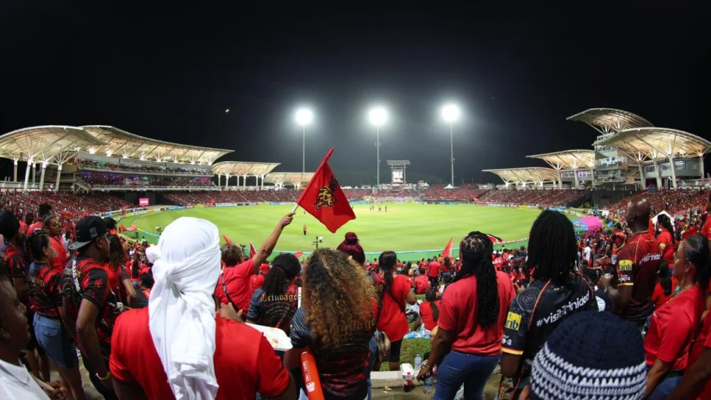 Packed stadium with energetic crowd dressed in red during a Caribbean Premier League (CPL) 2025 night match in Trinidad, fans waving flags and cheering under floodlights.