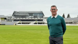 Jon Lewis standing on a cricket field in a teal sweater with a stadium pavilion in the background