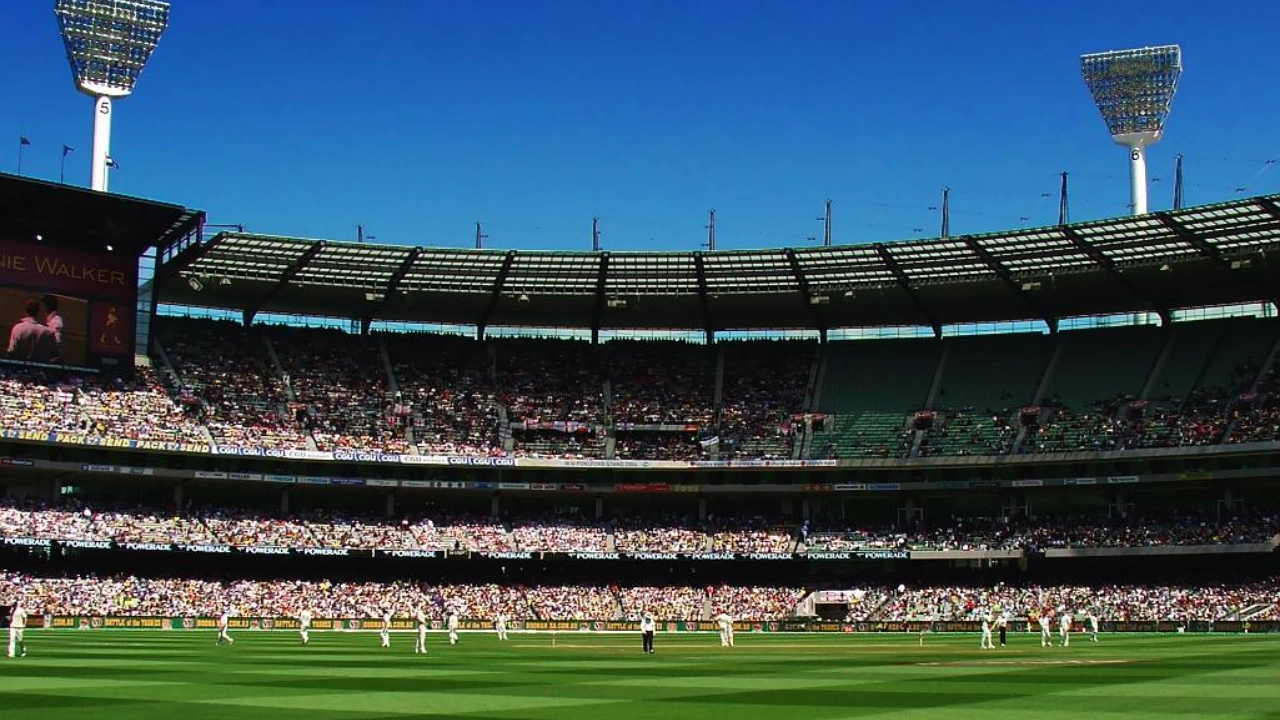 Wide-angle view of a cricket match at Melbourne Cricket Ground with a packed crowd under a clear blue sky.