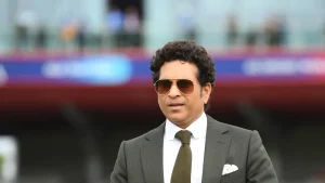 Sachin Tendulkar in a suit and sunglasses at a cricket stadium, with a blurred crowd in the background.