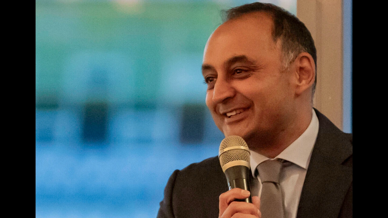 Cricket executive Sanjay Patel speaking at an event with a microphone in hand and smiling