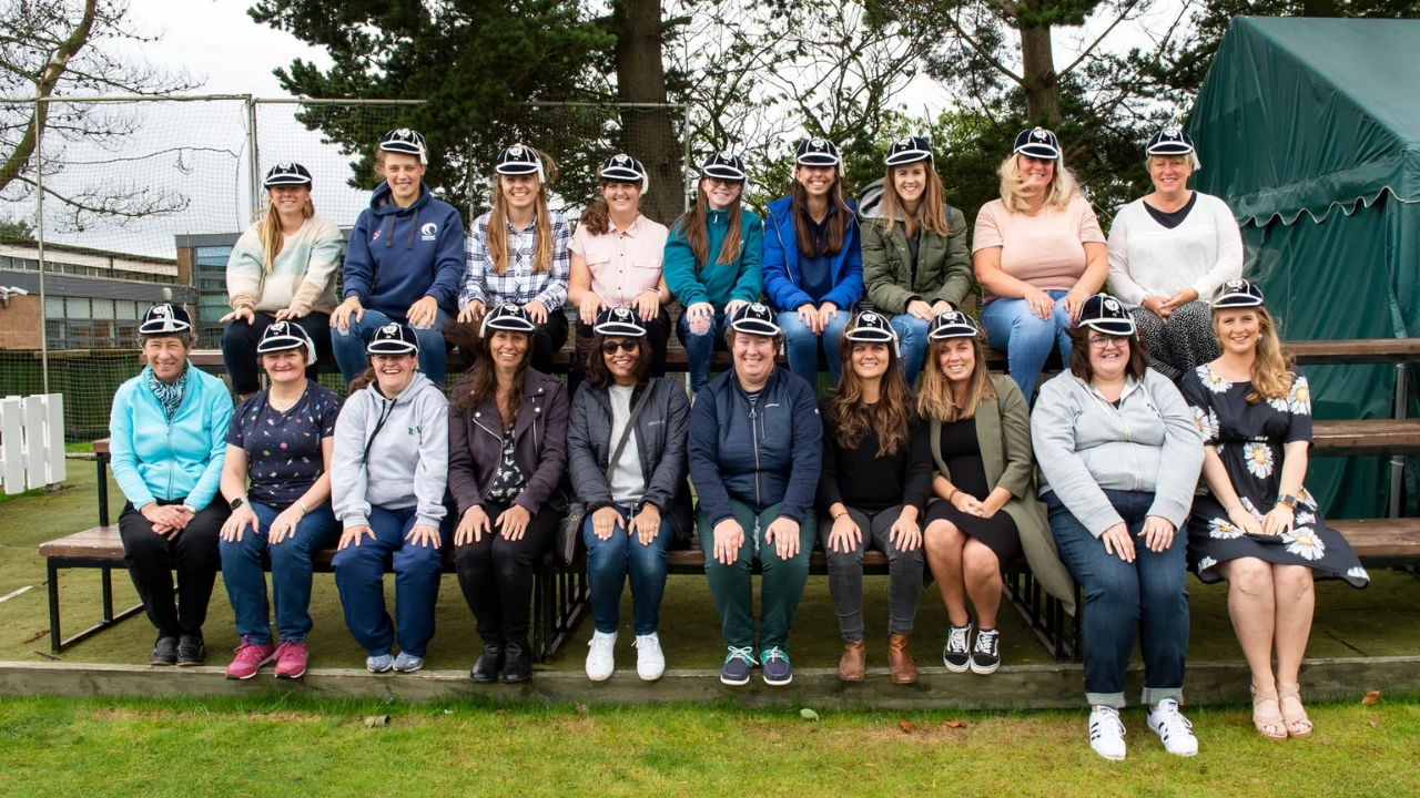 Group photo of Scotland women's cricket alumni wearing commemorative caps, seated on bleachers at a cricket ground in 2025.