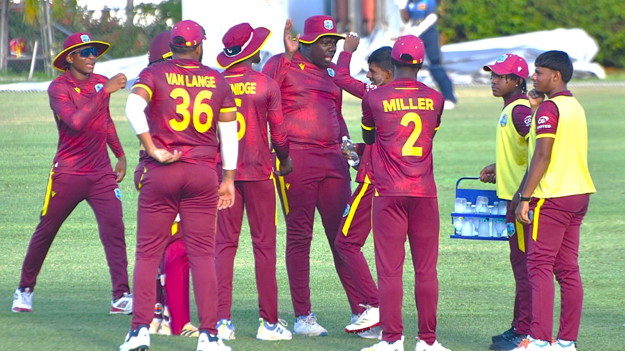 West Indies Under-19 cricket team players gathered on the field during a match, wearing maroon kits with yellow accents.