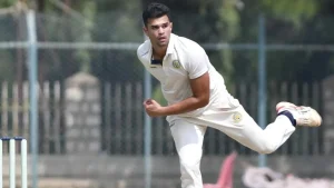 Arjun Tendulkar in action during an Indian domestic cricket match, captured mid-delivery in white cricket kit with stumps visible in the foreground.