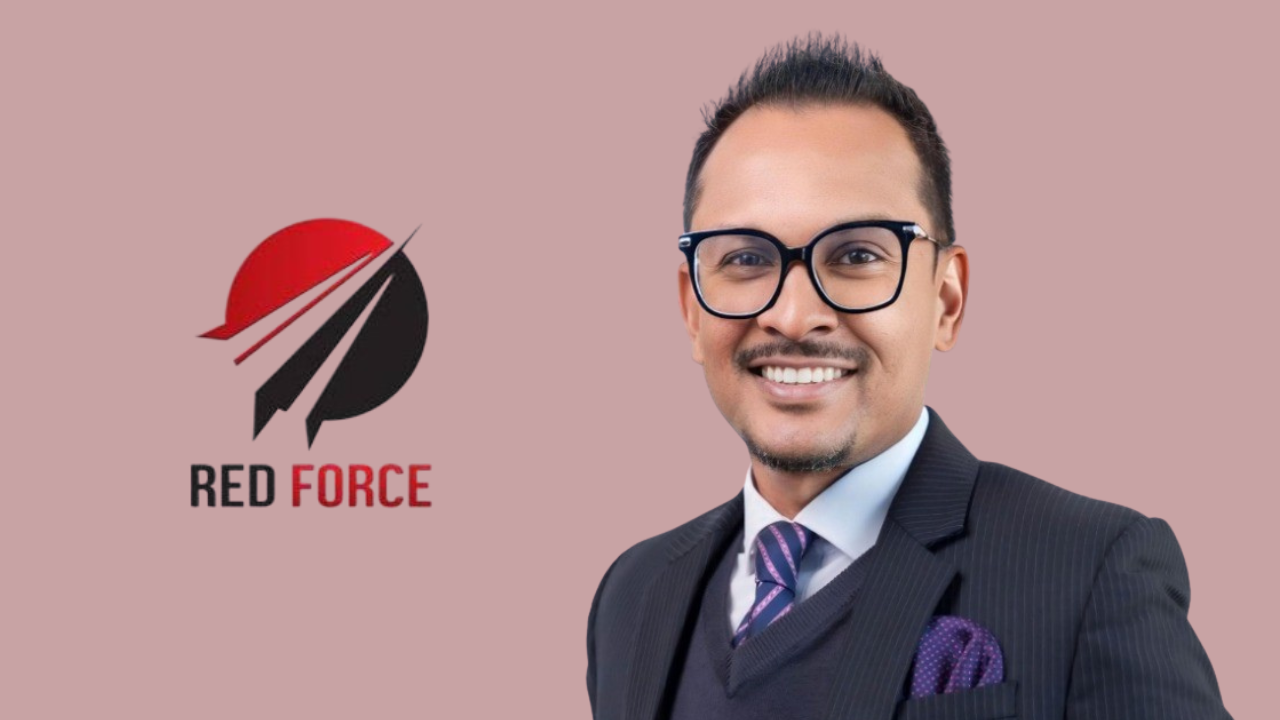 Avinash Suruj in formal attire standing next to the TTCB Red Force cricket team logo