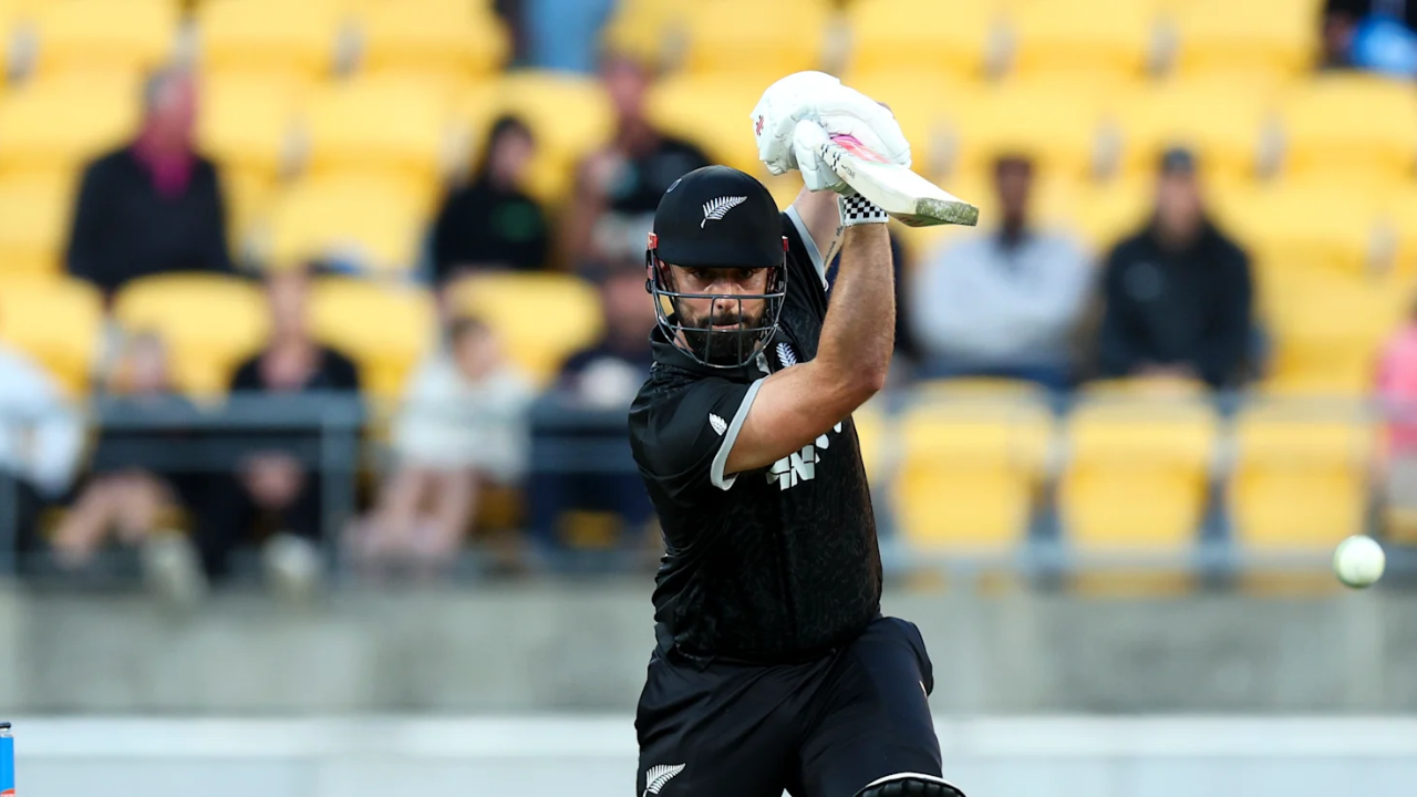 New Zealand cricketer Daryl Mitchell in action, playing a shot during a match in front of a crowd in a stadium with yellow seats.