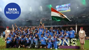 Indian women’s cricket team and support staff celebrating with the trophy under the Indian flag after a championship win, with Tata Motors logo visible in the background.