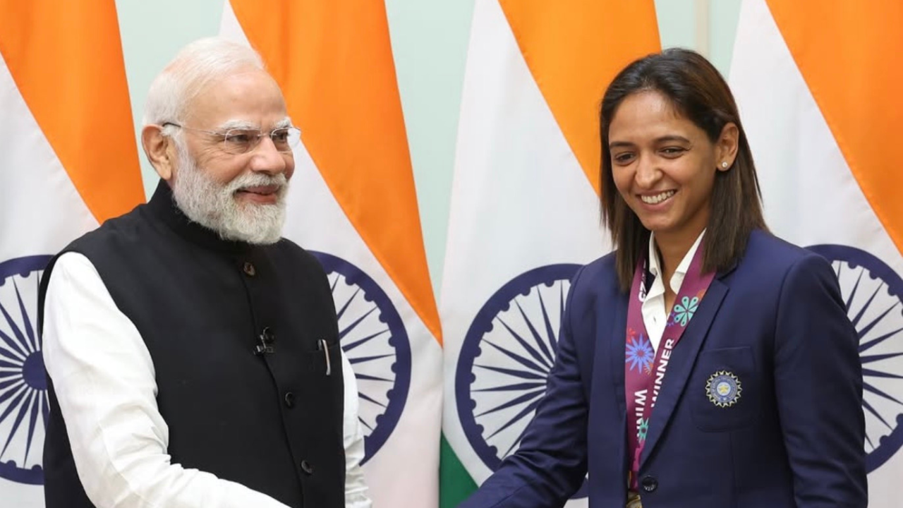 Indian Prime Minister Narendra Modi shaking hands with women’s cricket team captain Harmanpreet Kaur, who is wearing a medal and team blazer, in front of Indian flags.