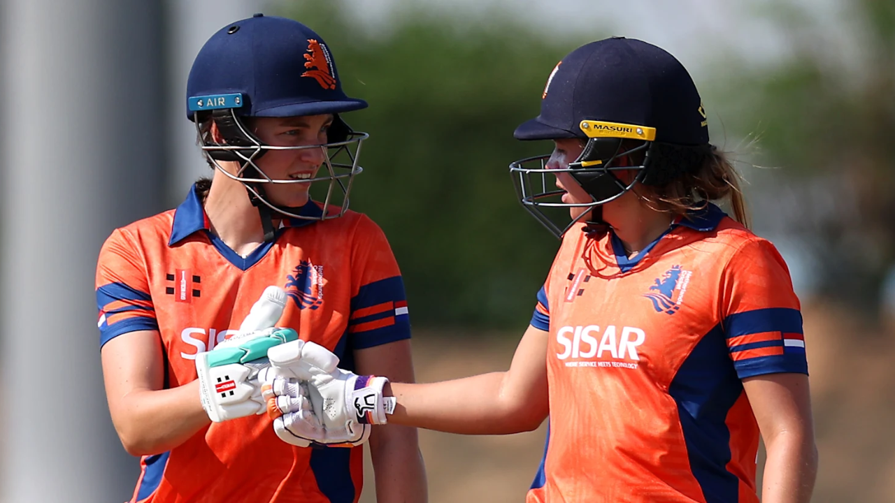 Two women cricketers from Netherlands team celebrating batting partnership during a match, wearing orange jerseys