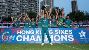 Team Pakistan celebrates winning the Hong Kong Sixes 2025, holding the trophy and standing behind a champions banner on the field with city buildings in the background.