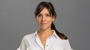 Professional headshot of Ros Trinick wearing a white shirt and gold jewelry against a gray background