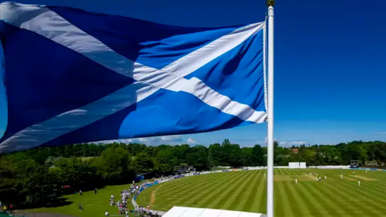 Scottish flag waving above a cricket ground during a match on a sunny day