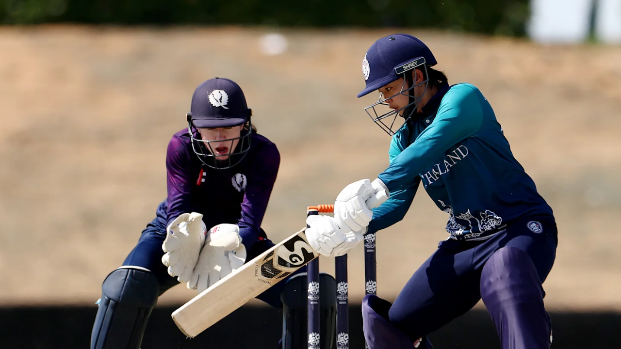 Thailand women's cricketer plays a shot while Scotland wicketkeeper watches closely during an international match, both in full playing kits.