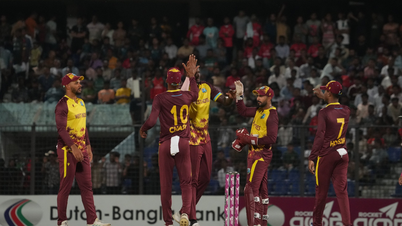 West Indies cricket team players celebrate a wicket during a T20 match, wearing maroon and yellow uniforms with a cheering crowd in the background