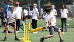 Young boy playing a cricket shot during a school cricket match on a turf field, with teammates and coach watching in the background.