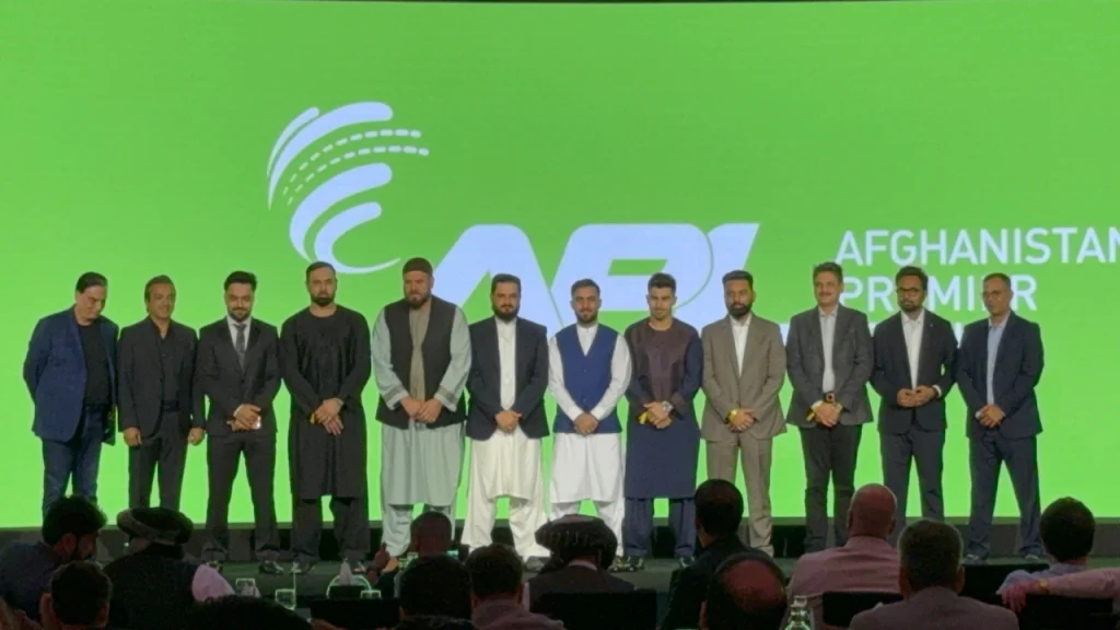Executives and star Afghan cricketers, including Rashid Khan and Mohammad Nabi, posing together on stage during the Afghanistan Premier League 2025 launch event, with APL branding on a green background.
