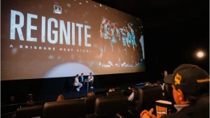 Audience watching the premiere of "Reignite: A Brisbane Heat Story" documentary in a cinema, with a panel discussion taking place in front of the large screen.