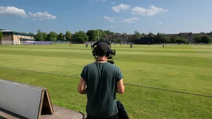 Camera operator recording a live cricket match on a sunny day from the boundary line