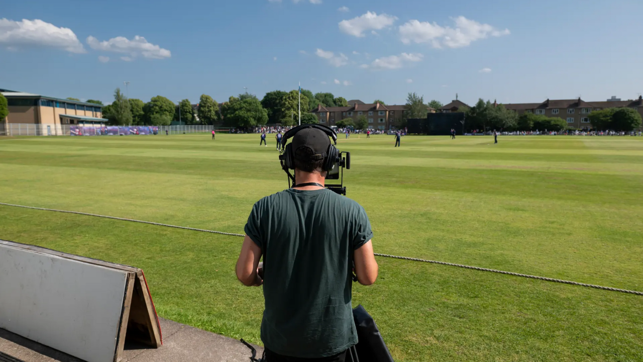 Camera operator recording a live cricket match on a sunny day from the boundary line