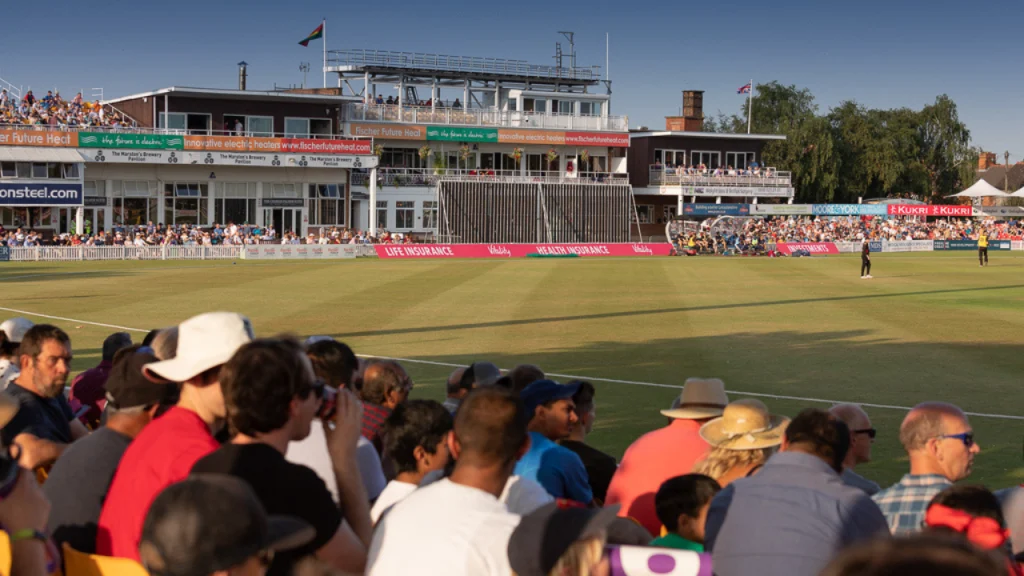 Crowd watching a live county cricket match at a sunlit stadium with a full stand and player activity on the field.