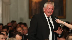 David Graveney smiling while speaking into a microphone at a formal event, wearing a black suit and tie