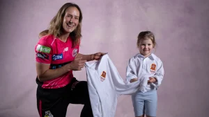 Ellyse Perry smiling with a young girl while holding an Oakhill College cricket shirt during a promotional photoshoot.