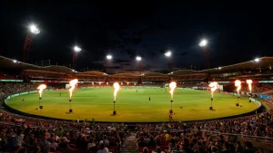 Night view of a cricket match at ENGIE Stadium with crowd, bright floodlights, and flame effects around the field