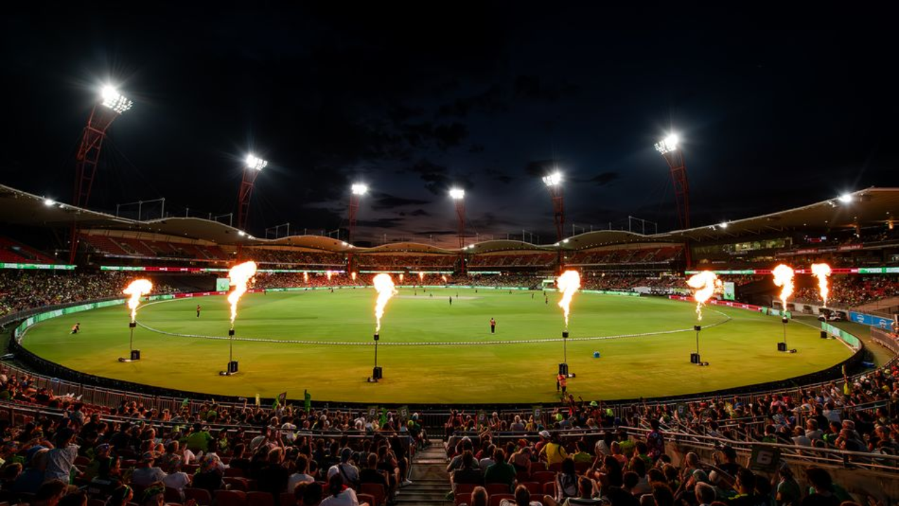 Night view of a cricket match at ENGIE Stadium with crowd, bright floodlights, and flame effects around the field