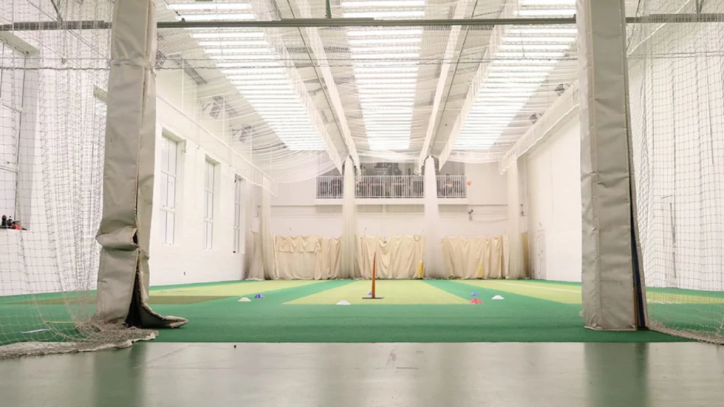 Indoor cricket practice facility with training nets, stumps, and colored cones under bright lighting.