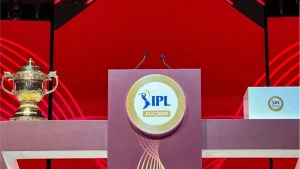 IPL auction rostrum with microphones flanked by the IPL trophy on a red stage background