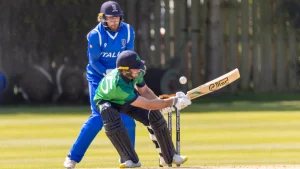 Cricket Ireland batter playing a shot while Cricket Italy wicketkeeper watches during a live match, with both players in full cricket gear on a sunny day.