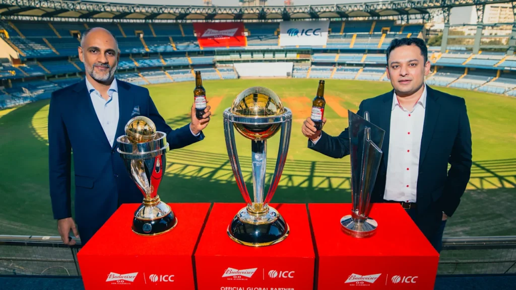 AB InBev CMO Marcel Marcondes and ICC CEO Sanjog Gupta holding Budweiser bottles next to ICC trophies at stadium partnership event