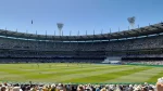 Panoramic view of the Melbourne Cricket Ground (MCG) during a match, showing the Members' Reserve from the Shane Warne Stand with a large crowd and players on the field under a clear blue sky.