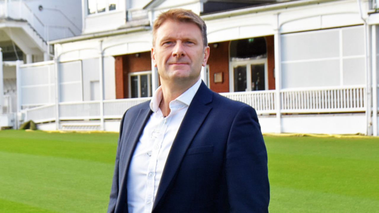 Michael Wilshaw standing on a cricket ground in a formal suit, with pavilion in the background