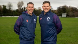 Cricket Scotland coaches Owen Dawkins and Gordon Drummond posing together on a cricket field in team training gear.