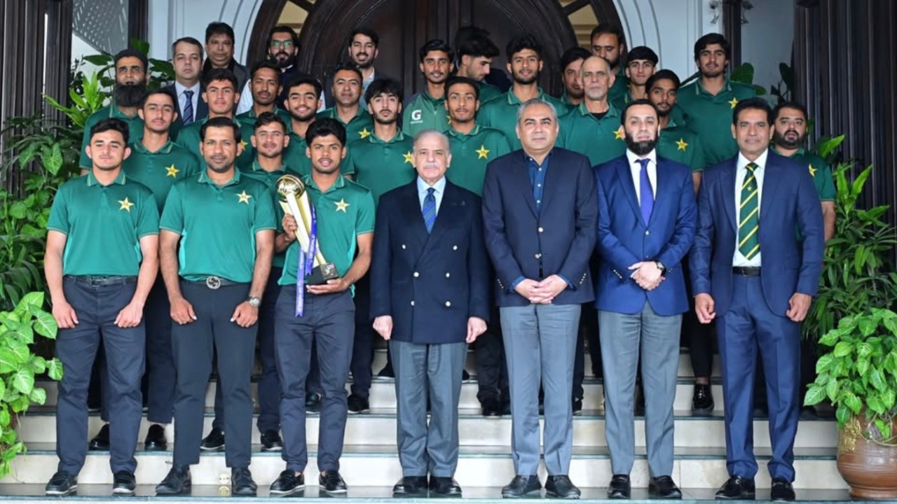 Prime Minister Shahbaz Sharif, Mohsin Naqvi, Attaullah Tarrar, and Aaqib Javed posing with the Pakistan U19 cricket team holding a trophy on the steps of an official building.
