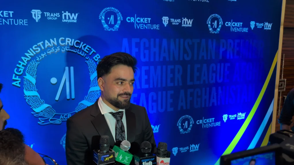 Rashid Khan wearing a black suit and tie speaking to media at the launch ceremony of the Afghanistan Premier League, with official event backdrop and microphones in front.