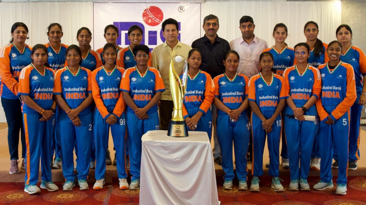 Sachin Tendulkar with India's women's blind cricket team posing with a trophy at an indoor event supported by IndusInd Bank