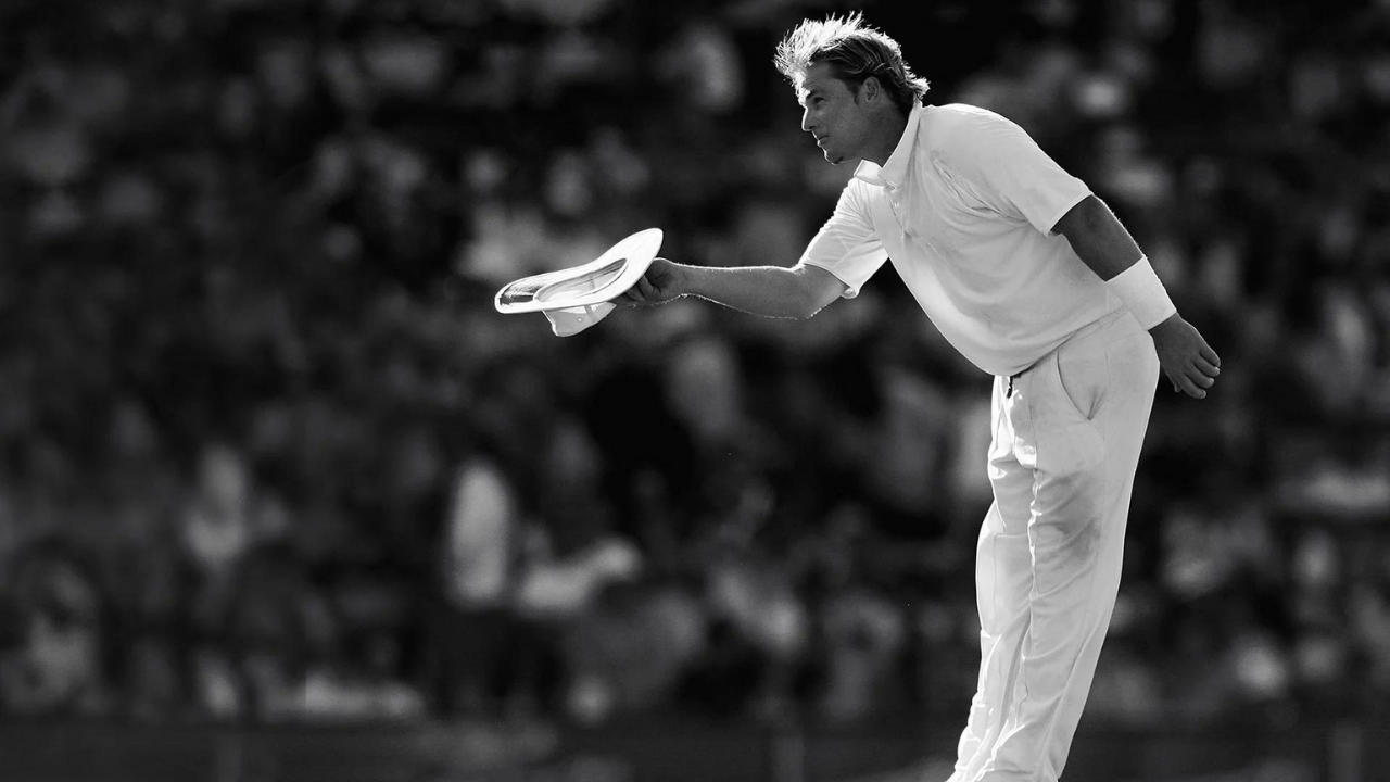 Shane Warne acknowledges the crowd during a cricket match, captured in a black and white tribute photograph.