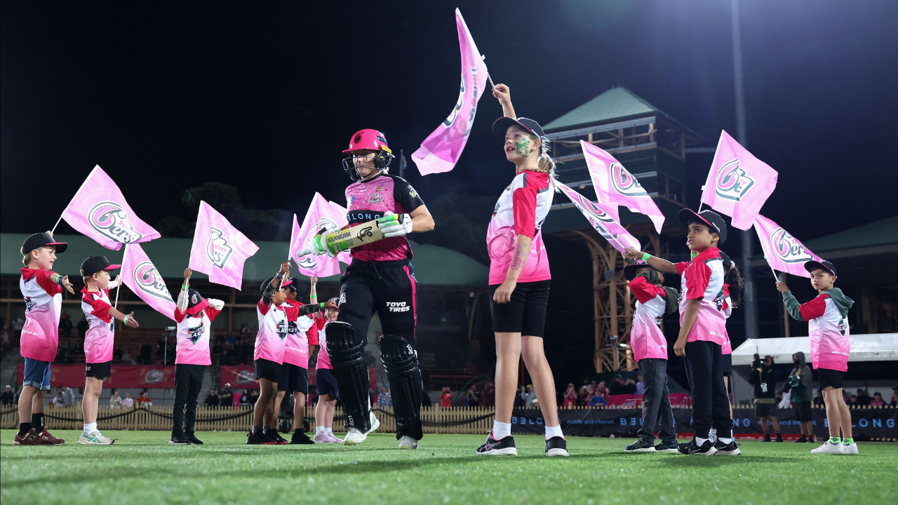 Alyssa Healy walks out to bat wearing the Sydney Sixers jersey, greeted by children waving Sixers flags during a BBL match.