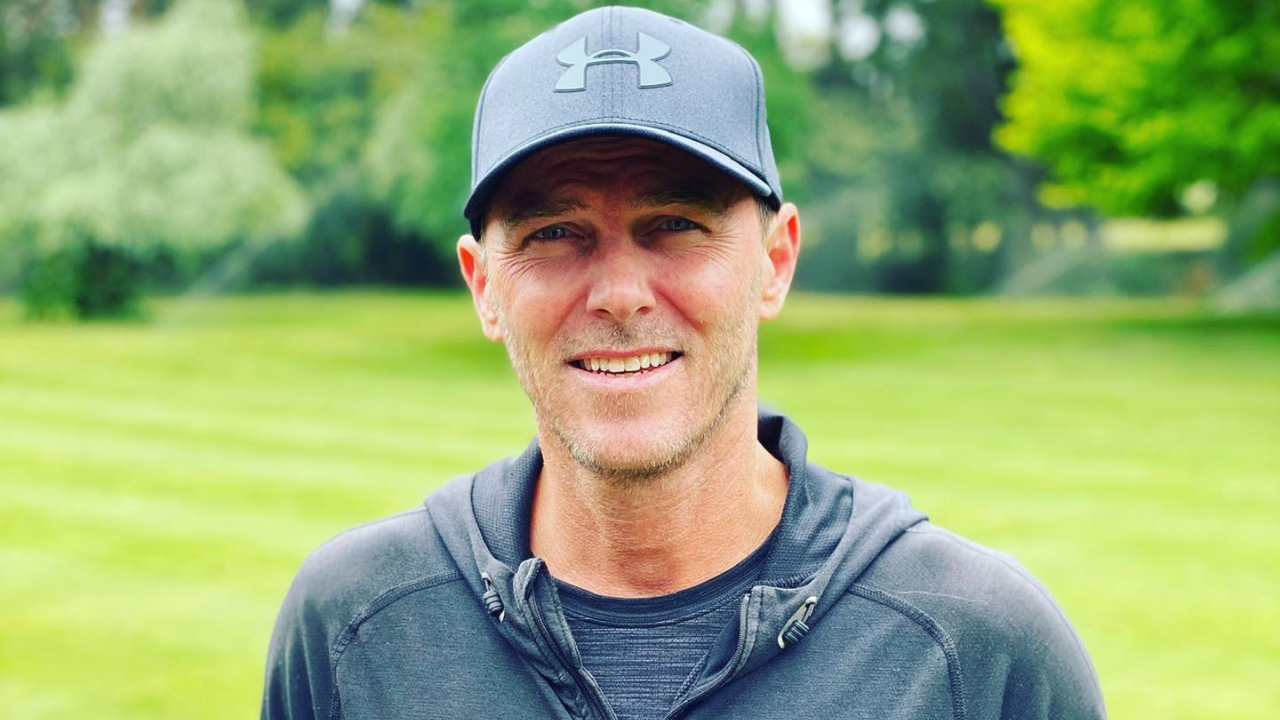 Former cricketer Damien Martyn wearing a cap and hoodie, smiling outdoors with a green grassy background
