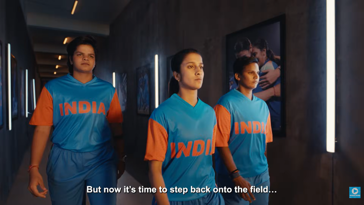 Three Indian women cricketers in blue and orange uniforms walking confidently through a stadium tunnel