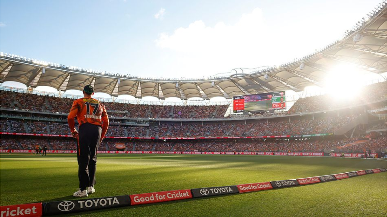 Ashton Turner of the Perth Scorchers stands on the boundary during a Big Bash League match at Optus Stadium in Perth, with a large crowd and sunset in the background.