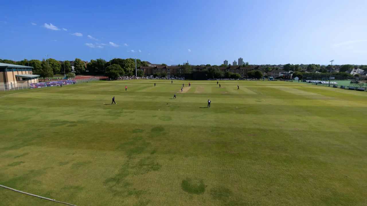Wide-angle view of a cricket match being played on a sunny day at a local ground in England, with spectators in the background and a clear blue sky overhead.