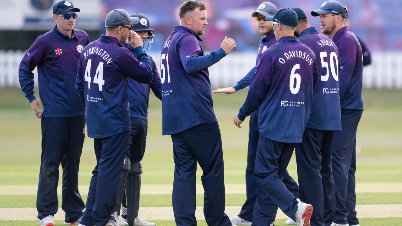 Scotland men's cricket team players in navy and purple kits celebrating on the field during a match, with visible names including Berrington, Watt, Sharif, and O Davidson.