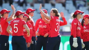 England women’s cricket players celebrate a wicket during an international match, wearing red kits on the field.