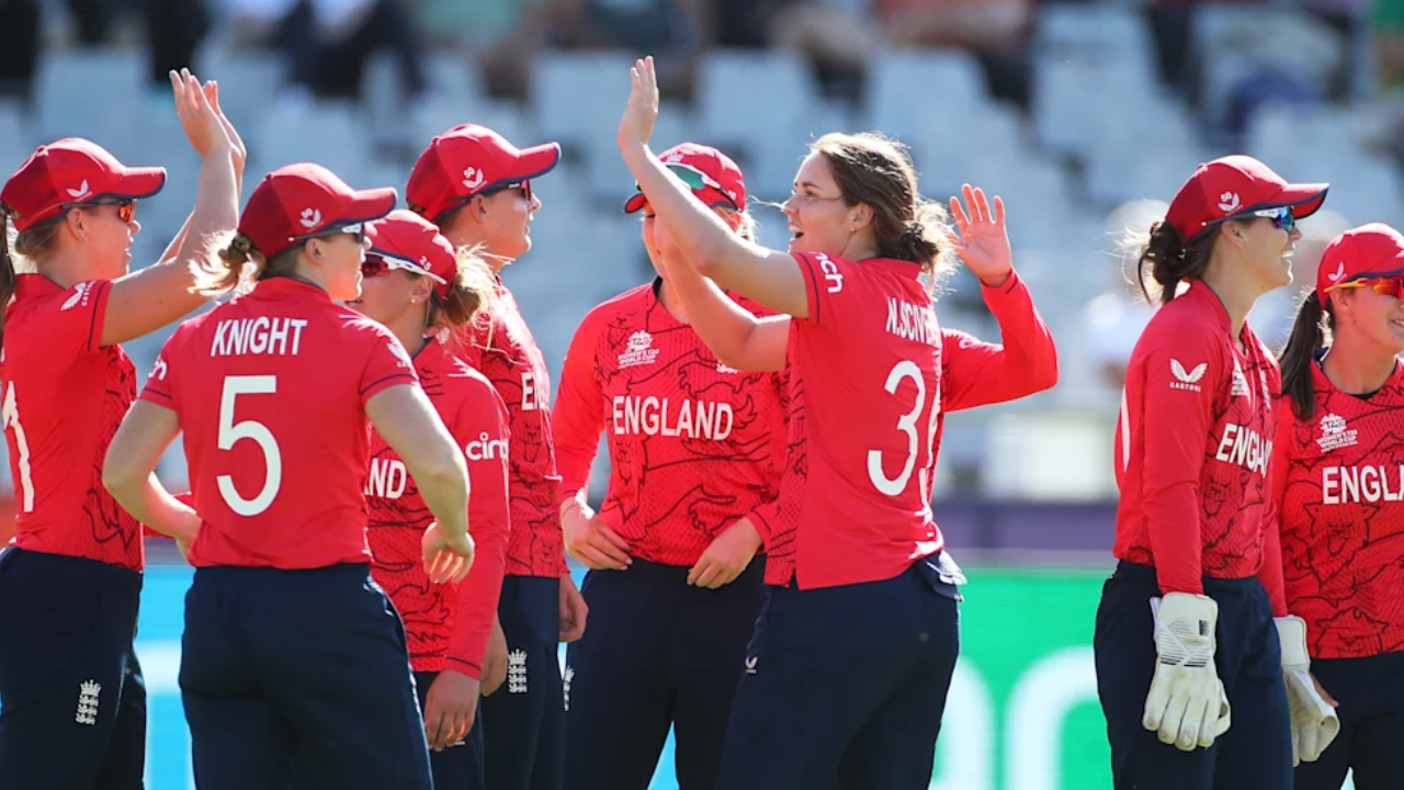 England women’s cricket players celebrate a wicket during an international match, wearing red kits on the field.