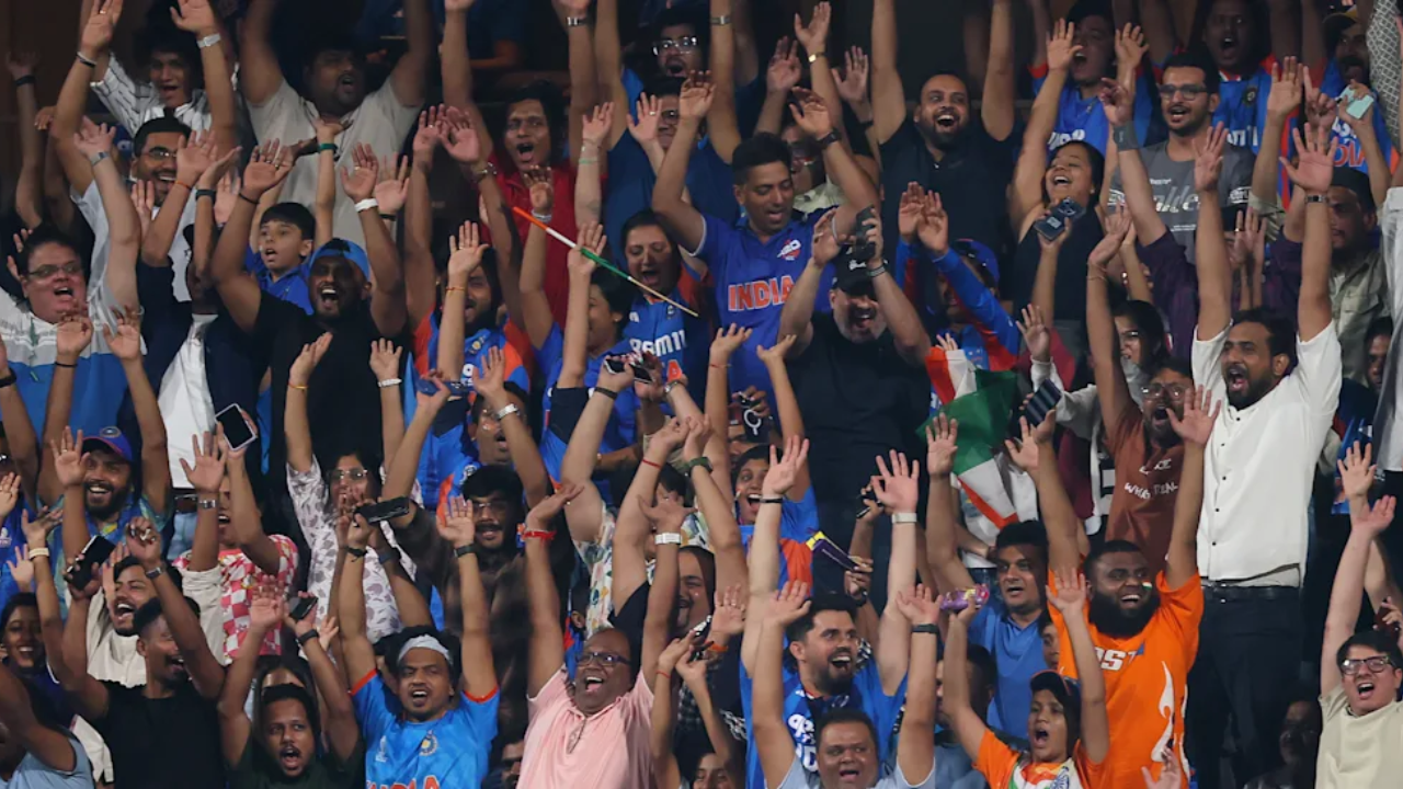 Indian cricket fans celebrating with raised hands in a packed stadium during an international match, many wearing Team India jerseys and waving the national flag.